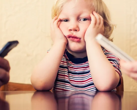 A young child in a striped shirt sits at a table with hands on cheeks, looking upset as two adults hold phones nearby.