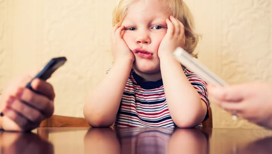 A young child in a striped shirt sits at a table with hands on cheeks, looking upset as two adults hold phones nearby.