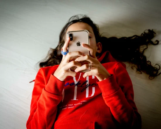 Person lying on a light floor in a red hoodie, holding an iPhone up to their face to use the device.