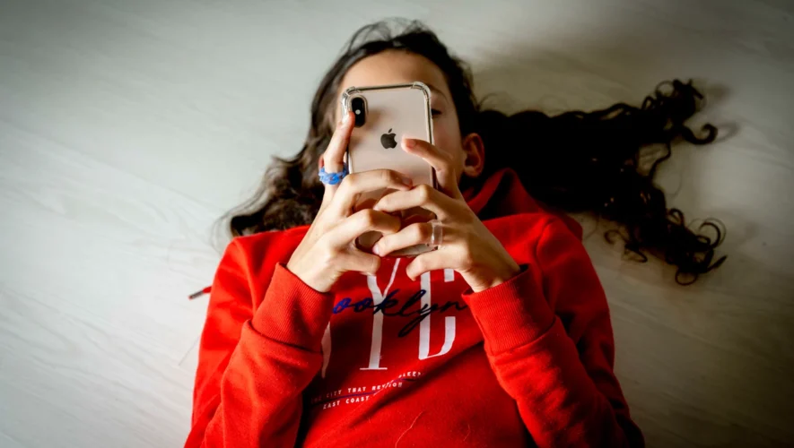 Person lying on a light floor in a red hoodie, holding an iPhone up to their face to use the device.