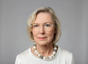Portrait of an older woman with short blonde hair and glasses, wearing a white top and beaded necklace against a gray backdrop.