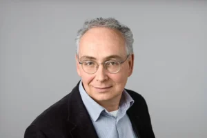 Professional headshot of a middle-aged man with gray hair and glasses, wearing a light blue shirt and dark blazer against a neutral gray background.