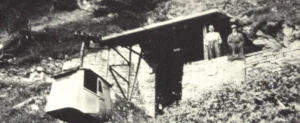 Old black-and-white photo of a small shed built into a rocky hillside, with two people standing near the top right area.",