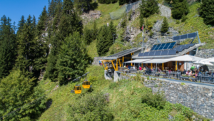 Mountain restaurant perched on a hillside with a yellow cable car gliding by, solar panels on the roof, and outdoor dining set among trees.