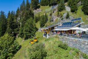 Mountain restaurant perched on a hillside with a yellow cable car gliding by, solar panels on the roof, and outdoor dining set among trees.