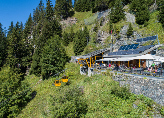 Mountain restaurant perched on a hillside with a yellow cable car gliding by, solar panels on the roof, and outdoor dining set among trees.