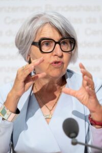 Older woman with gray hair and black glasses speaking at a podium, gesturing with her hands in a conference setting.