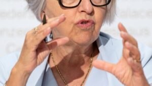 Older woman with gray hair and black glasses speaking at a podium, gesturing with her hands in a conference setting.