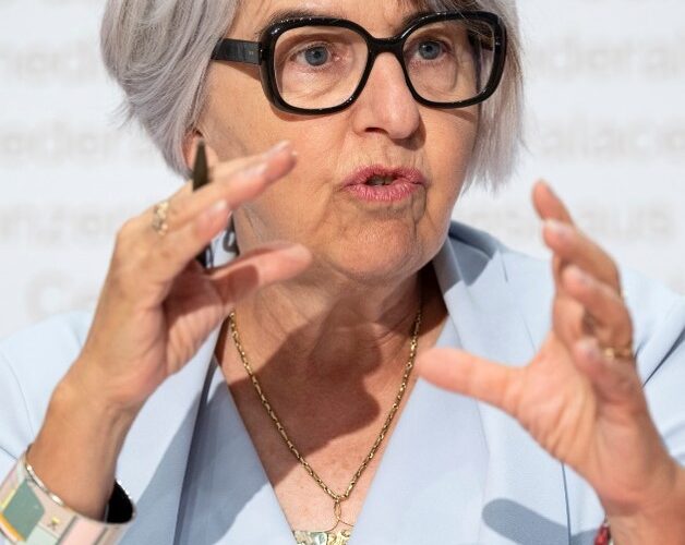 Older woman with gray hair and black glasses speaking at a podium, gesturing with her hands in a conference setting.
