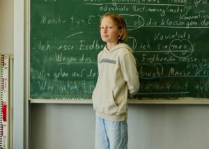 Young student in a beige hoodie stands at a chalkboard covered with German writing in a classroom.