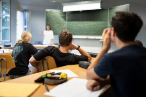 Students in a classroom listening to a teacher at the chalkboard.