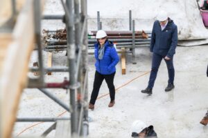 Two construction workers wearing hard hats walk across a concrete site with scaffolding and pipes in the background.