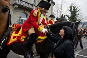 Woman on the street shakes hands with a man in a bright red historical uniform riding a dark horse during a parade on a city street.