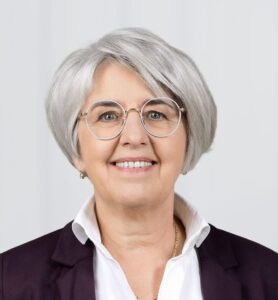 Professional headshot of a smiling older woman with short gray hair, round glasses, white shirt, and navy blazer.