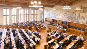 Parliament chamber with rows of wooden desks, lawmakers at work, a large mural along the back wall and hanging chandeliers above.