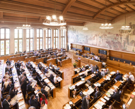 Parliament chamber with rows of wooden desks, lawmakers at work, a large mural along the back wall and hanging chandeliers above.