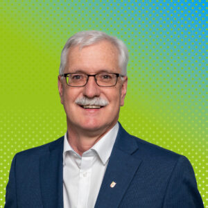 Portrait of an older man with white hair and glasses, wearing a blue suit, smiling against a lime-green, cross-patterned background.