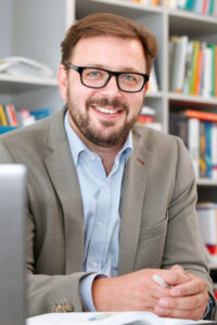 Smiling man wearing glasses and a blazer sits at a desk in a bookshelf-filled office, holding a pen.