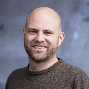 Portrait of a smiling bald man with a short beard wearing a brown sweater against a blue-gray blurred background, head-and-shoulders shot.