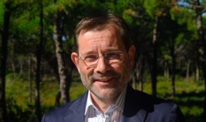 Portrait of a middle-aged man with short brown hair and glasses, smiling outdoors in a sunlit forest, wearing a navy blazer and striped shirt.