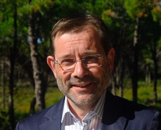 Portrait of a middle-aged man with short brown hair and glasses, smiling outdoors in a sunlit forest, wearing a navy blazer and striped shirt.