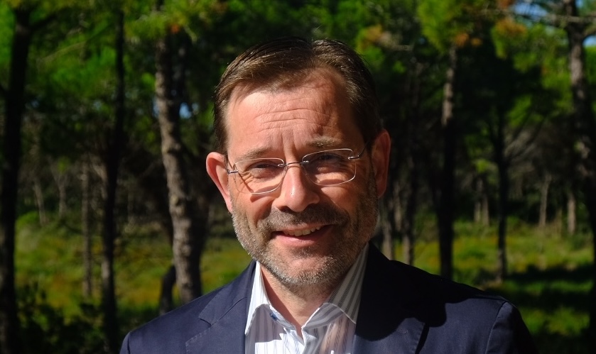 Portrait of a middle-aged man with short brown hair and glasses, smiling outdoors in a sunlit forest, wearing a navy blazer and striped shirt.