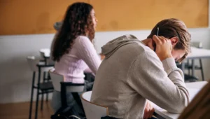 Student in a gray hoodie rests his head on his arm while holding a pencil at a classroom desk.