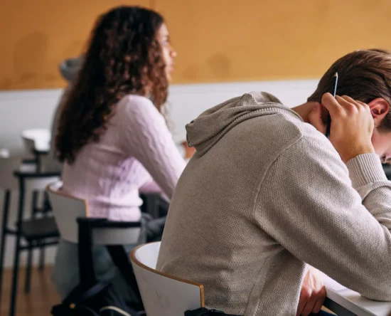 Student in a gray hoodie rests his head on his arm while holding a pencil at a classroom desk.