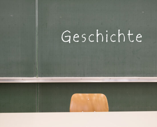 Green chalkboard in a classroom with 'Geschichte' written in white chalk and a wooden chair in front of it.
