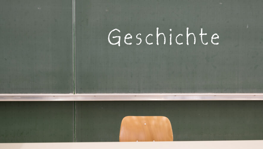 Green chalkboard in a classroom with 'Geschichte' written in white chalk and a wooden chair in front of it.