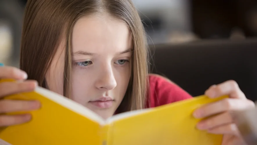 Young girl with long hair focused on reading a bright yellow book.