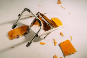 Collapsed folding chair with orange seat and gray metal frame on a light floor, with wood shavings scattered around.