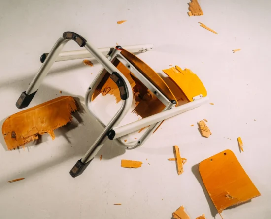 Collapsed folding chair with orange seat and gray metal frame on a light floor, with wood shavings scattered around.