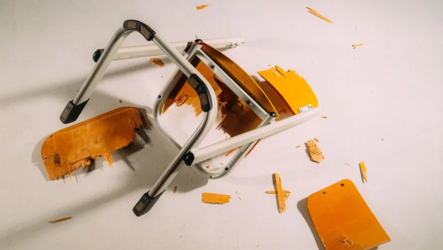Collapsed folding chair with orange seat and gray metal frame on a light floor, with wood shavings scattered around.