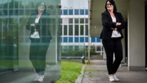 Professional woman with glasses in a black blazer and white lace top stands with arms crossed beside a glass building; her reflection is visible in the glass.