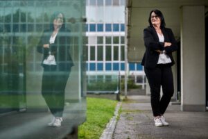 Professional woman with glasses in a black blazer and white lace top stands with arms crossed beside a glass building; her reflection is visible in the glass.