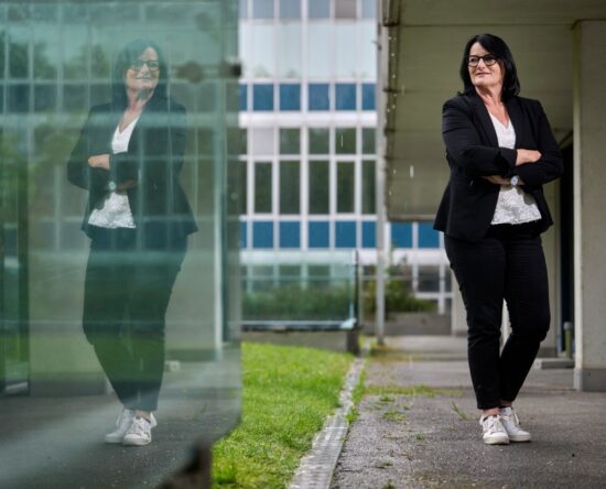 Professional woman with glasses in a black blazer and white lace top stands with arms crossed beside a glass building; her reflection is visible in the glass.