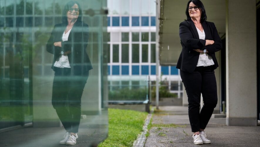 Professional woman with glasses in a black blazer and white lace top stands with arms crossed beside a glass building; her reflection is visible in the glass.