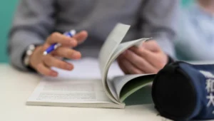 Hands of a person writing with a blue pen while flipping through a small booklet on a desk surface.