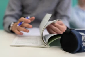 Hands of a person writing with a blue pen while flipping through a small booklet on a desk surface.
