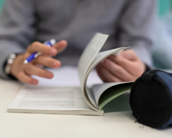 Hands of a person writing with a blue pen while flipping through a small booklet on a desk surface.