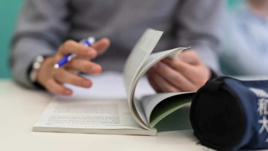 Hands of a person writing with a blue pen while flipping through a small booklet on a desk surface.