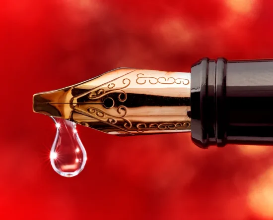 Close-up of a gold fountain pen nib with a droplet of ink hanging from its tip against a red blurred background.