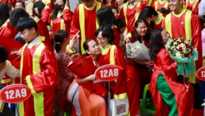 Crowded graduation scene with students in red gowns and yellow sashes; some graduates hug, hold flowers, and signs like 12A8 and 12A9 are visible as families cheer.