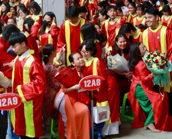 Crowded graduation scene with students in red gowns and yellow sashes; some graduates hug, hold flowers, and signs like 12A8 and 12A9 are visible as families cheer.