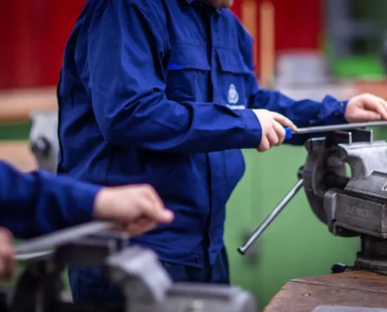 Two workers in blue overalls sharpening metal on a bench vise in a workshop with a red background behind them.