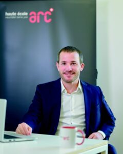 Portrait of a man in a navy blazer at a desk, with a laptop and mug, in front of a backdrop showing arc logo.
