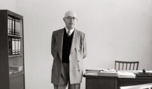Older man in a double-breasted suit stands with hands behind his back in a simple office, with a desk and filing cabinet nearby.