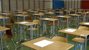 Empty school gym set up with rows of desks and papers for an exam or event, wooden chairs, and gym mats in the background, green floor.