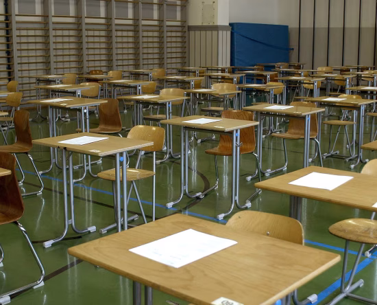 Empty school gym set up with rows of desks and papers for an exam or event, wooden chairs, and gym mats in the background, green floor.